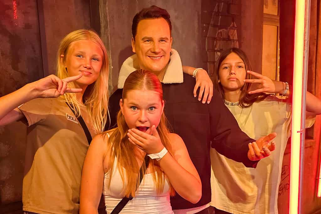 Drei gut gelaunte Personen einer Jugendgruppe posieren mit V-Zeichen und überraschten Gesten vor rotem Licht bei Madame Tussauds in Berlin.
